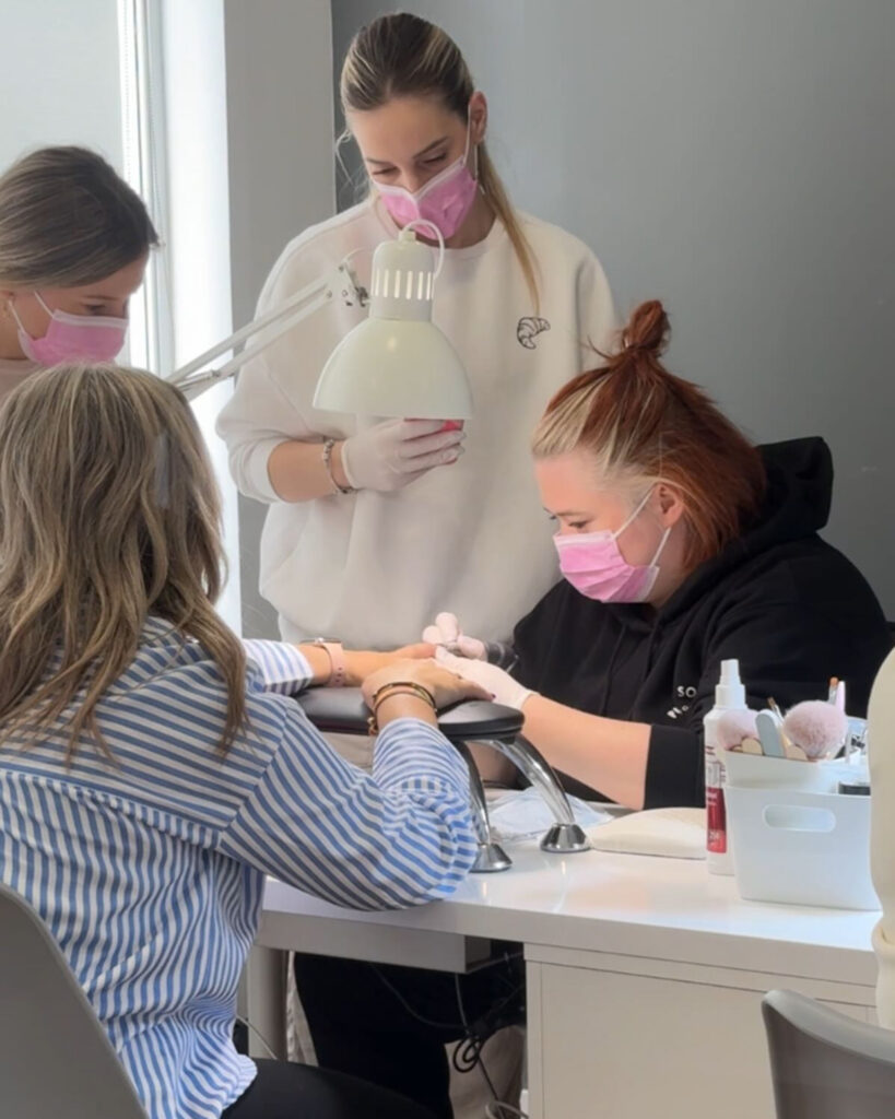 Katysol de Katysol Nails realizando una manicura mientras es observada por dos asistentes o estudiantes, todas llevando guantes y mascarillas protectoras de color rosa, demostrando higiene en el salón.