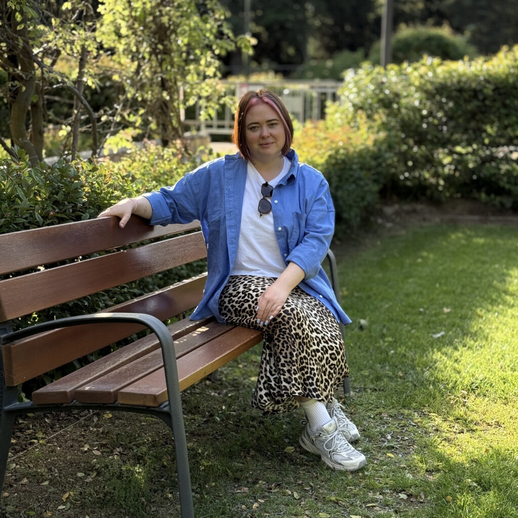 Katysol de Katysol Nails sentada en un banco de parque bajo el sol, vestida con ropa casual (camisa azul y falda de leopardo)