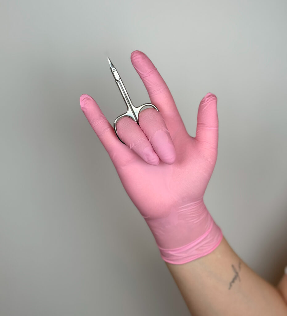 Mano enguantada de Katysol con guante rosa sosteniendo unas tijeras de manicura profesionales para el corte de cutículas.